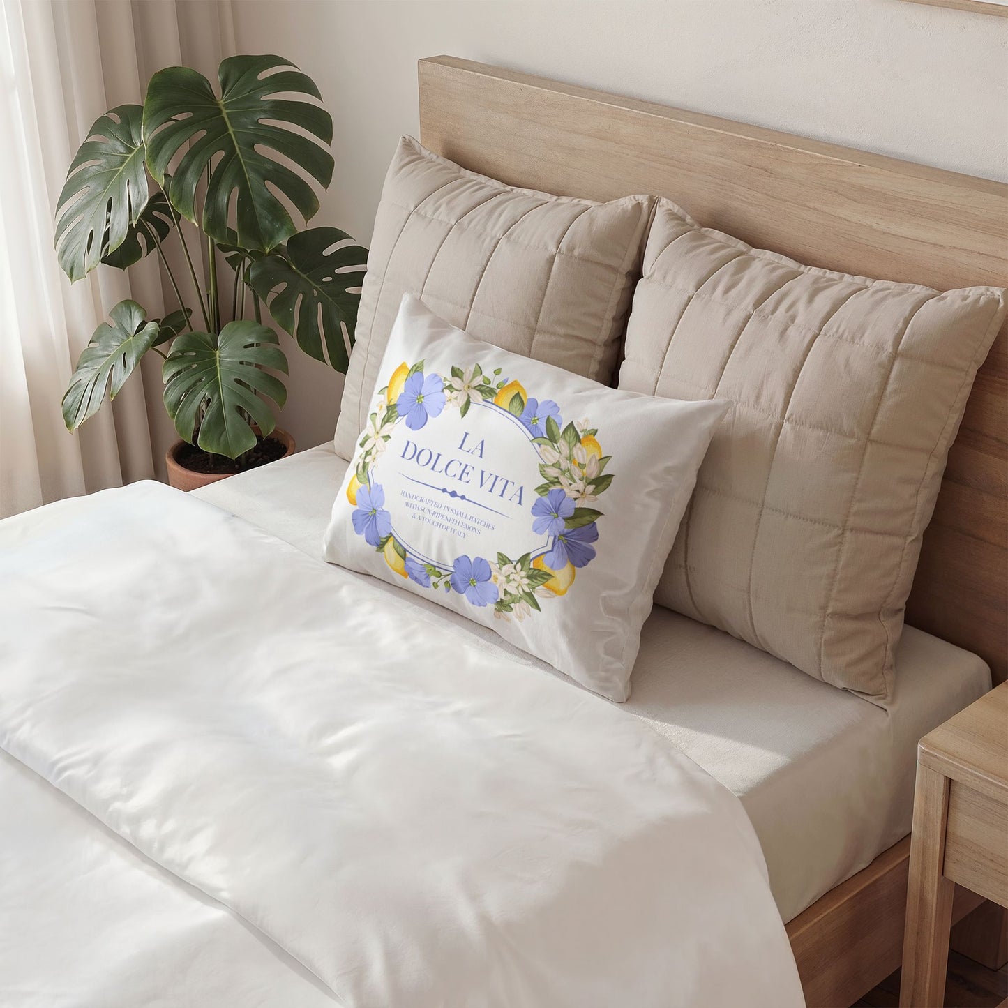 White cotton pillow sham with lemon and violet floral wreath design featuring La Dolce Vita text, displayed on a made bed beside a monstera plant