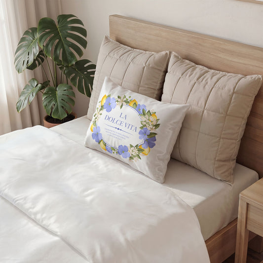 White cotton pillow sham with lemon and violet floral wreath design featuring La Dolce Vita text, displayed on a made bed beside a monstera plant
