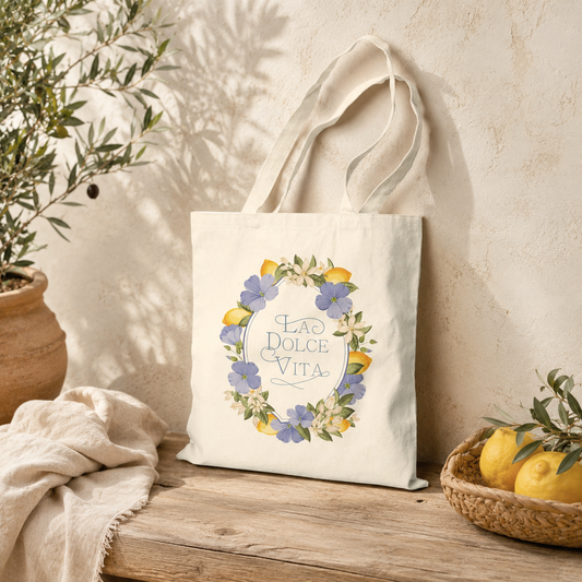 Natural cotton canvas tote bag with a lemon and violet floral wreath design resting on a wooden bench beside fresh lemons and an olive plant