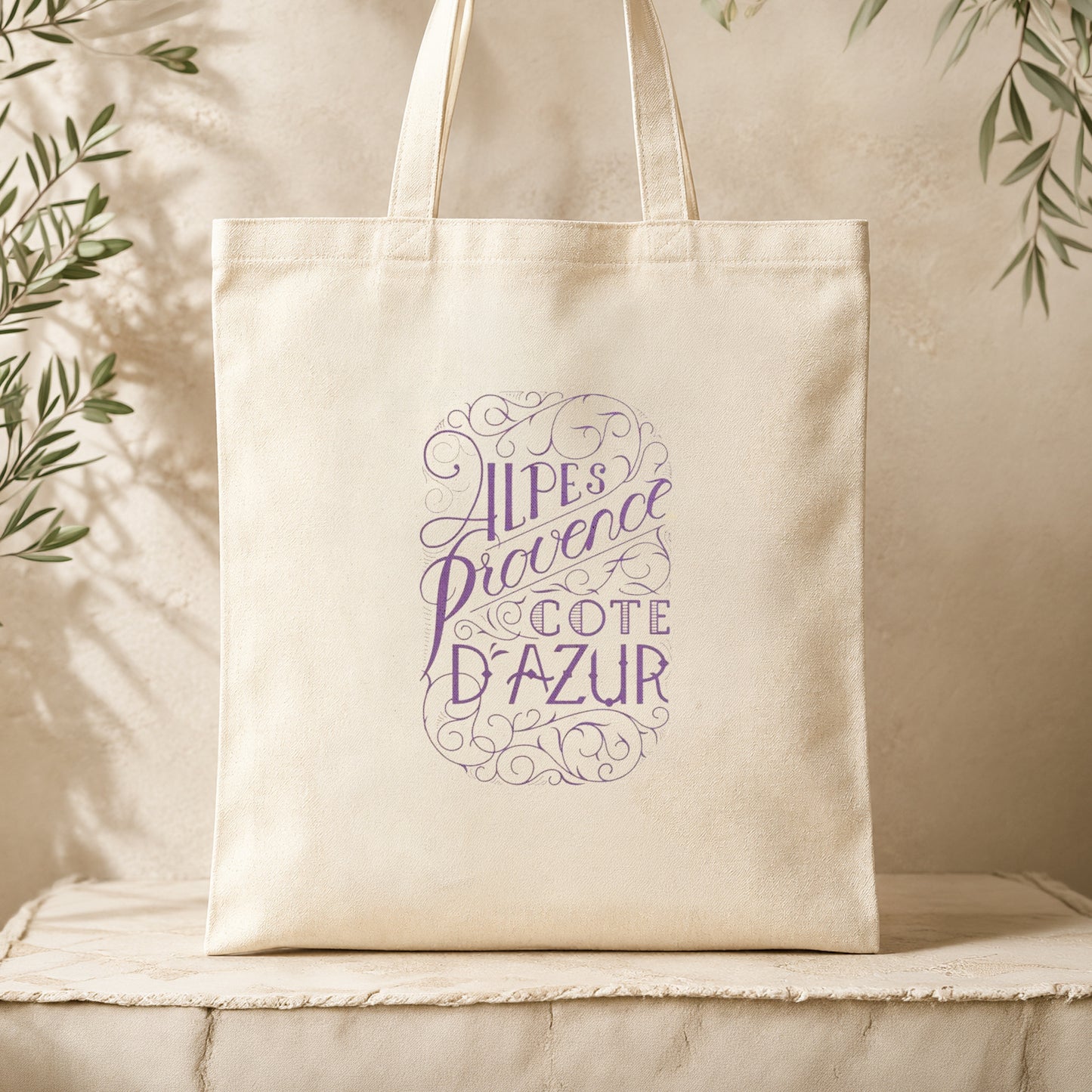 Natural canvas tote bag with lavender inscription resting on a cream textured linen surface surrounded by dried olive branches