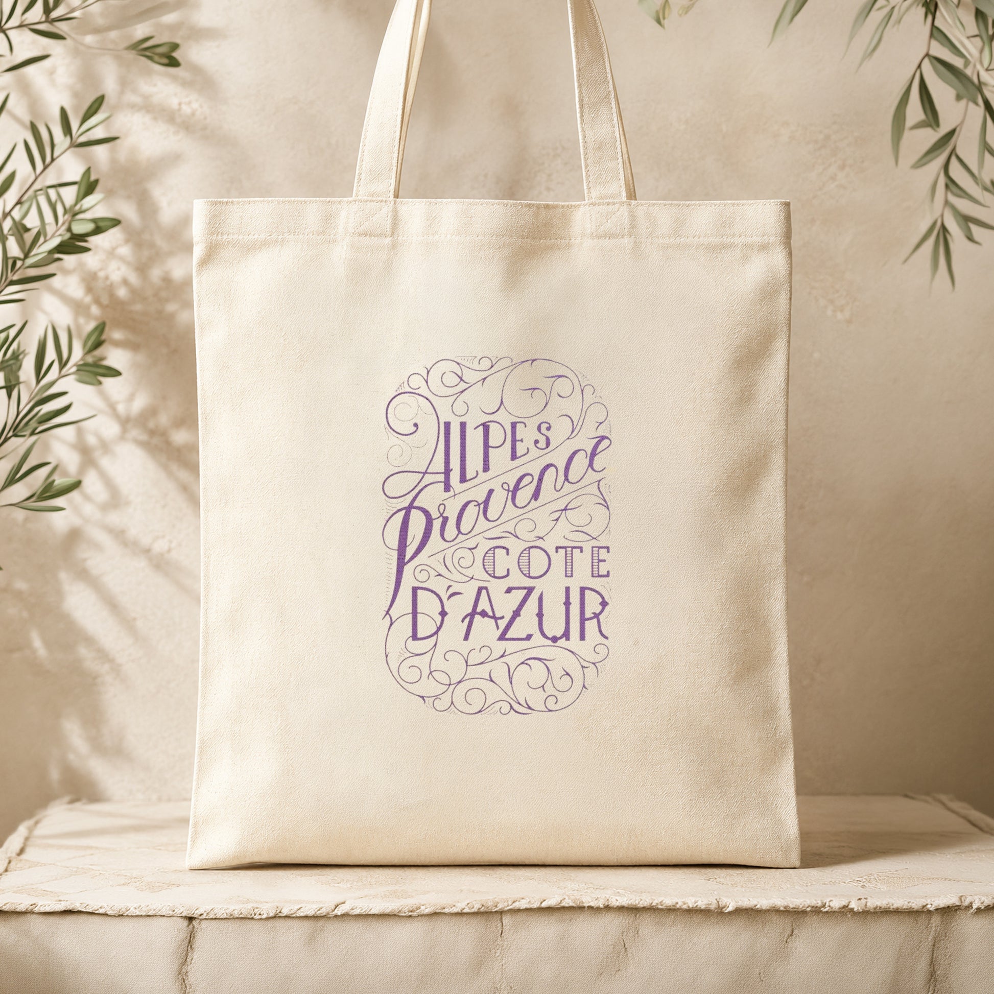 Natural canvas tote bag with lavender inscription resting on a cream textured linen surface surrounded by dried olive branches