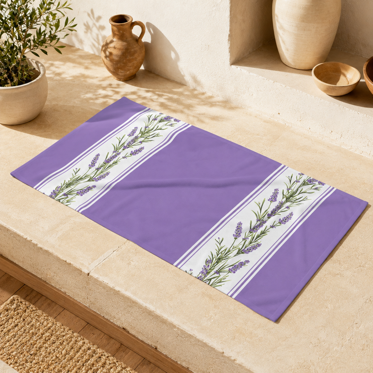 Lavender stripe beach towel folded and displayed on a cream stone surface, styled with Mediterranean accessories including a potted herb plant, terracotta bowl, and wooden vessels in natural sunlight
