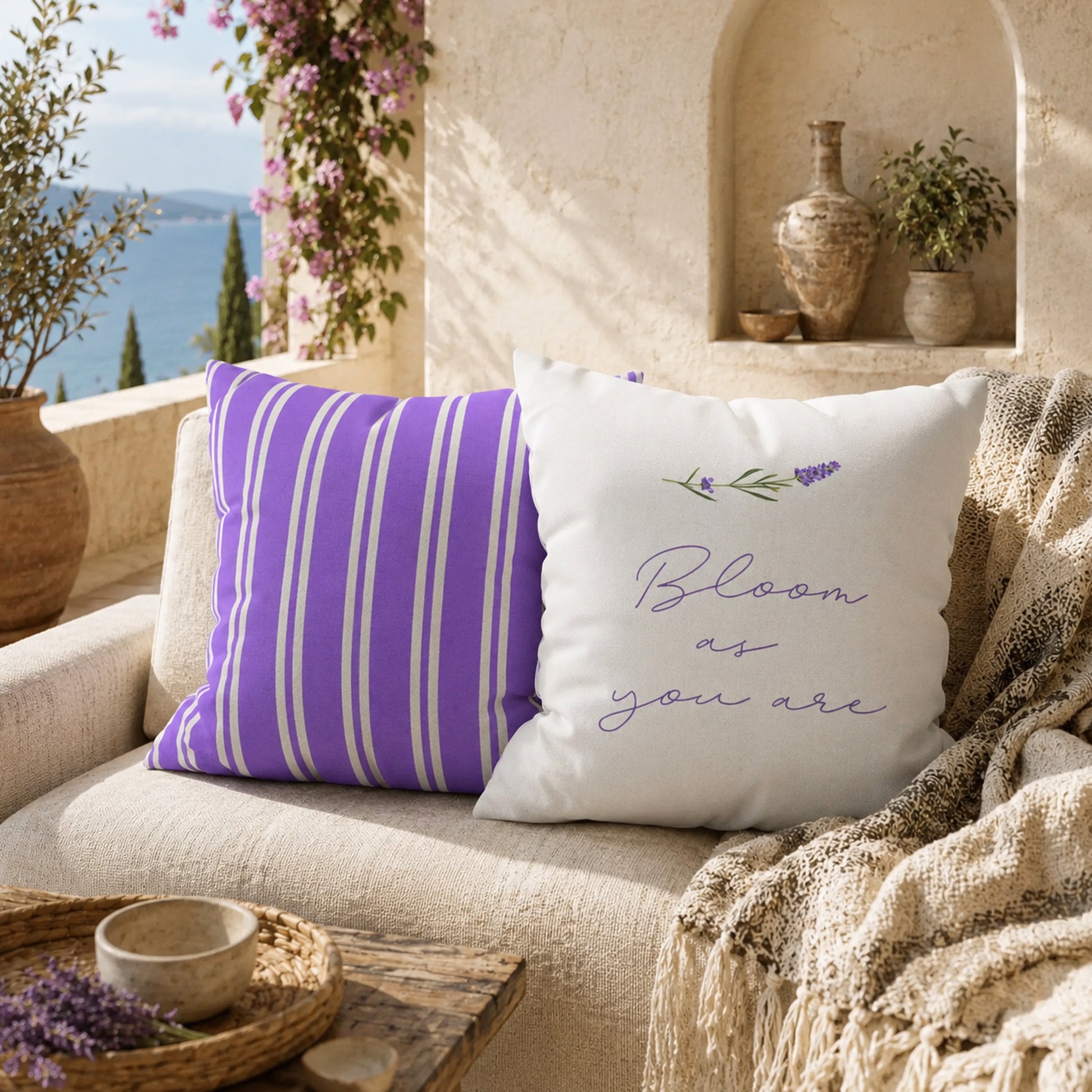 Lavender stripe and botanical text decorative pillows displayed on a cream outdoor sofa beside woven baskets and flowering vines on a Mediterranean patio